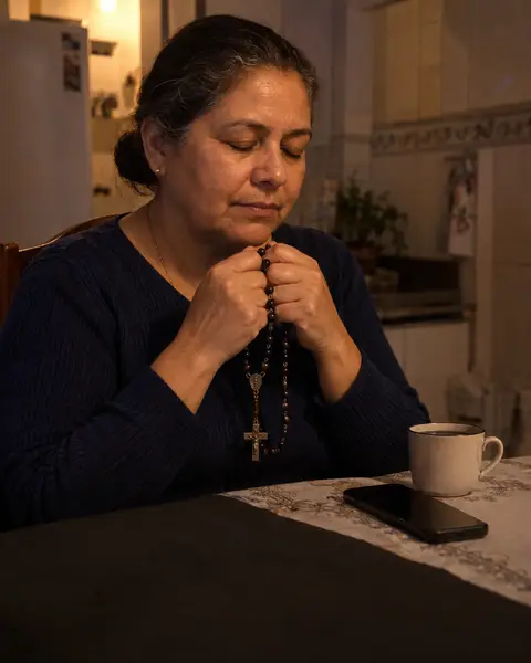 Mulher de meia-idade rezando com o terço nas mãos, à noite